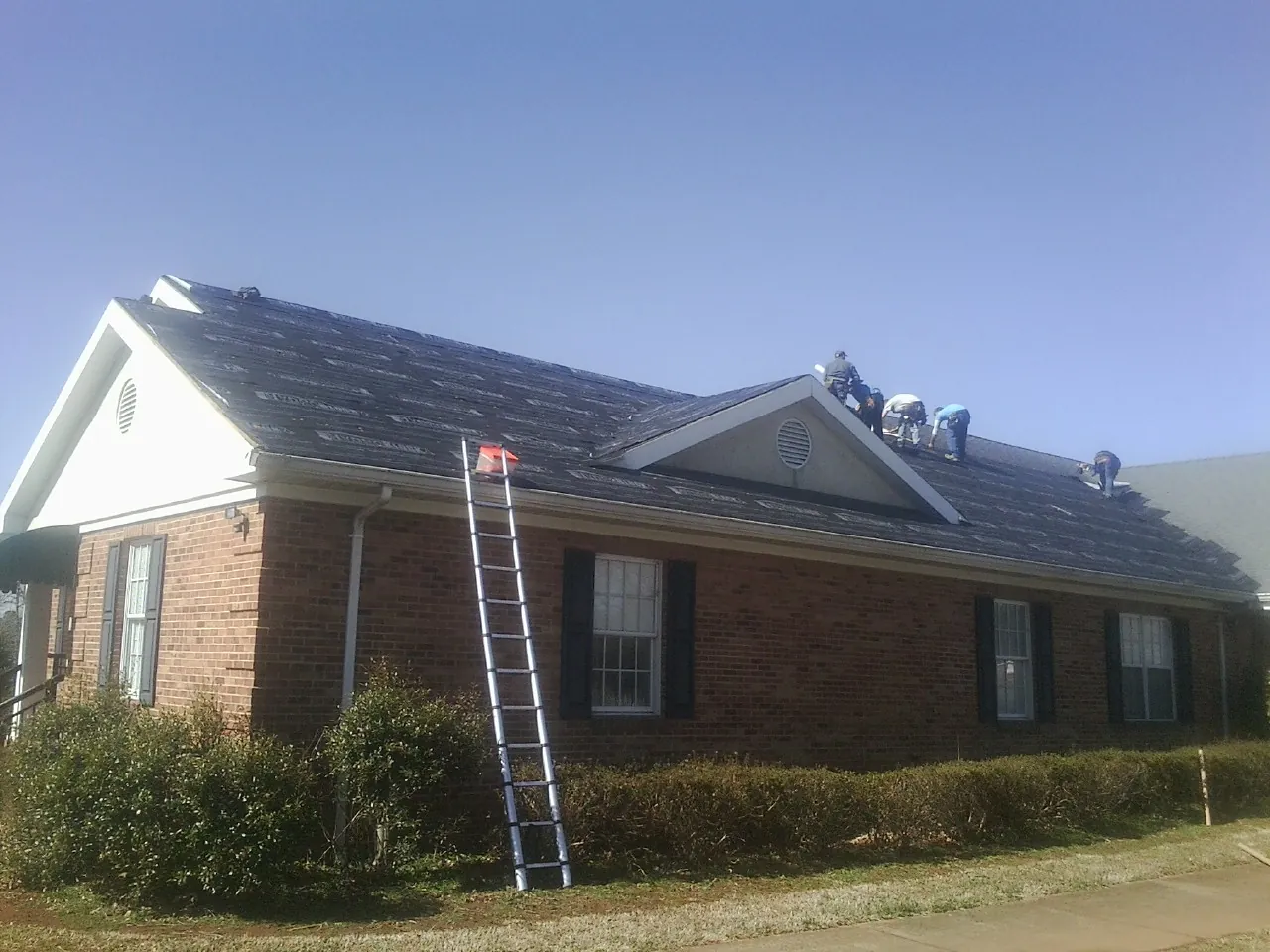Crew tearing shingles off a brick ranch roof with a ladder set — Emergency Roof Repair work by our crew in Grand Ledge