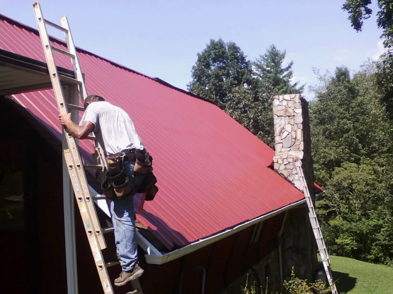 Roofer on a ladder beside a red metal roof and stone chimney — Soffit & Fascia Repair by Roofing Brothers in Grand Ledge